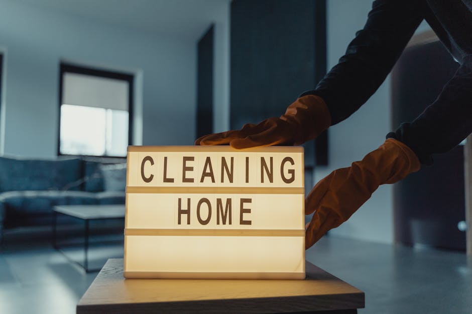 A person wearing brown cleaning gloves is adjusting a small illuminated sign that reads 'CLEANING HOME' on a wooden surface in a modern living room. The room features a dark-colored sofa with throw pillows, a coffee table, and large windows letting in natural light, highlighting the neat and dust-free surfaces. The space appears professionally cleaned, emphasizing hygiene and surface sanitation typical of deep domestic cleaning services offered by Tooting Carpet Cleaning near Tooting Bec Lido, with clean floors, polished surfaces, and organized furnishings.