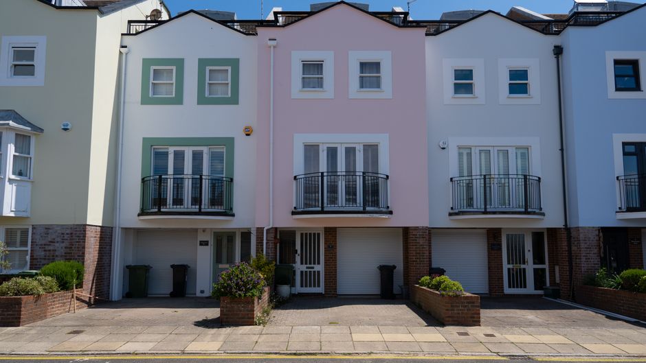 Photograph of a row of modern, multi-story residential buildings with pastel-colored facades, including shades of pink, light green, and white. The buildings feature small balconies with black metal railings, white-framed windows, and brick features at the base; some windows have shutters. The ground level has garage doors, with small paved driveways and neatly maintained garden beds with shrubs and flowers in front. Bright, natural daylight illuminates the scene, highlighting the tidy, well-kept appearance of the exterior. This image exemplifies the clean and orderly urban residential environment that Tooting Carpet Cleaning aims to maintain through its domestic cleaning and surface sanitisation services, especially near Tooting Bec Lido.