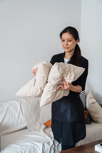 A woman with dark hair, dressed in a black uniform, stands in a brightly lit living room holding two off-white pillow covers or fabric items. The room features a white wall, a light-colored sofa with a beige cushion and an orange cushion on it, and a wooden table with some items partially visible. The sofa's surface appears clean and well-maintained, with no visible dust or stains. The overall setting suggests a tidy, modern interior, emphasizing domestic cleaning attention to detail, aligning with the upholstery cleaning options offered by Tooting Carpet Cleaning for local residents in Furzedown. Lighting is natural and evenly distributed, highlighting the cleanliness and arrangement of the space.
