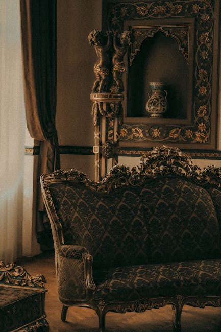 Interior view of an ornate living room featuring vintage-style furniture, including a dark upholstered sofa with intricate wood carvings and a matching armchair. The room has elaborate wallpaper with floral patterns and a large, decorative mirror framed with detailed carvings mounted on the wall. Beside the sofa, there is a tall, decorative pedestal with a classical-style vase. The space is illuminated by natural light coming through a window with dark curtains. The overall setting emphasizes a luxurious, antique aesthetic, with surfaces appearing well-maintained and clean, reflecting thorough domestic cleaning. This image exemplifies home decor that benefits from professional surface cleaning and maintenance, as provided by Tooting Carpet Cleaning, to ensure a hygienic and visually appealing environment.