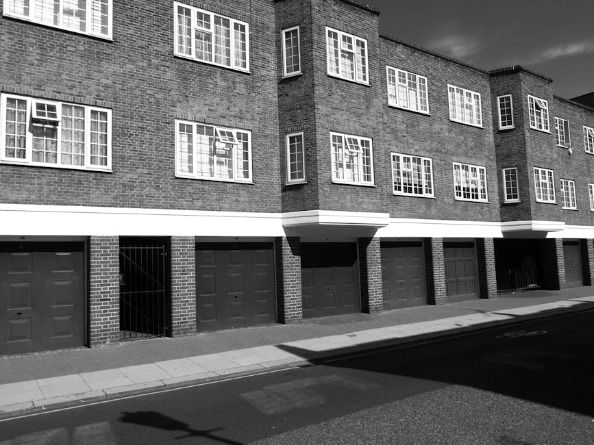 Black and white photograph of a modern residential building on Upper Tooting Road with multiple brick facades, featuring large rectangular windows with white frames, some open, and a series of garage doors at street level made of dark panels. The building has a clean, well-maintained appearance with smooth concrete walkways and a paved street in the foreground. Shadows cast by the overhanging structures indicate bright daylight. The image relates to property maintenance and could be associated with services provided by Tooting Carpet Cleaning in the context of thorough surface cleaning and sanitisation for residential flats.