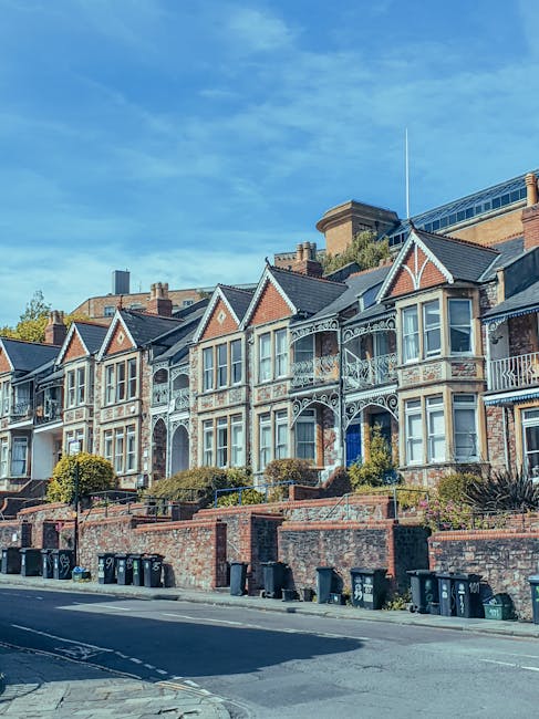 A row of Victorian-style terraced houses on Upper Tooting Road, featuring ornate white balconies and bay windows set on brick facades. The houses are positioned on a slight incline with front gardens bordered by low brick walls and neatly maintained shrubbery. The pavement and street in front are visible, with multiple black wheelie bins lined along the curbside. Bright daylight illuminates the scene, with a clear blue sky overhead. The image showcases an exterior residential setting, emphasizing the architectural details and well-kept appearance typical of domestic properties, as part of a content focus on surface cleaning and property maintenance. Tooting Carpet Cleaning is referenced as a professional service provider relevant to the setting.