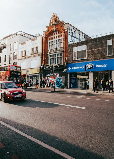 Exterior view of a commercial street in Tooting with a variety of storefronts, including a pharmacy and beauty shop, featuring large display windows and signage. The building facades combine brick and historic architectural elements, with the central building displaying a prominent arched window and decorative brickwork at the top. Pedestrians are walking along the sidewalk, some entering or exiting shops, and a red double-decker bus and a parked red car are visible on the street. The area is well-lit with natural daylight, and the scene captures a typical busy urban shopping area, emphasizing the importance of surface cleaning and maintenance for commercial properties. Tooting Carpet Cleaning, a professional cleaning service, specifically focuses on deep cleaning and sanitization of residential or commercial surfaces to maintain hygiene and cleanliness in such lively environments, aligning with the Tooting Broadway carpet cleaning guide for tenants.