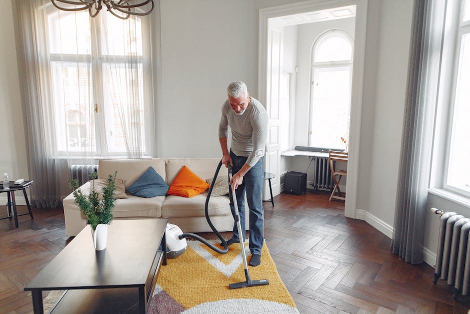A professional cleaner from Tooting Carpet Cleaning is performing surface cleaning in a bright, modern living room. He is using a vacuum cleaner on a light hardwood floor with a yellow and white geometric area rug. The room features large windows with sheer white curtains, allowing natural light to illuminate the space. There is a beige sofa with colorful cushions, a wooden coffee table with a potted plant, and a small side table near the window. The cleaner is dressed in casual attire and appears focused on deep cleaning the floor to ensure thorough hygiene and maintenance, reflecting domestic cleaning practices. The setting emphasizes a clean, well-maintained environment with attention to surface sanitisation and care of various materials, including wood floors and soft furnishings, under the expert service of Tooting Carpet Cleaning.